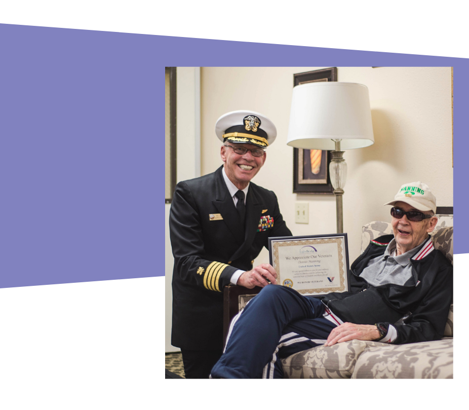 A man in uniform proudly holds a certificate beside a woman seated in a chair, both smiling.