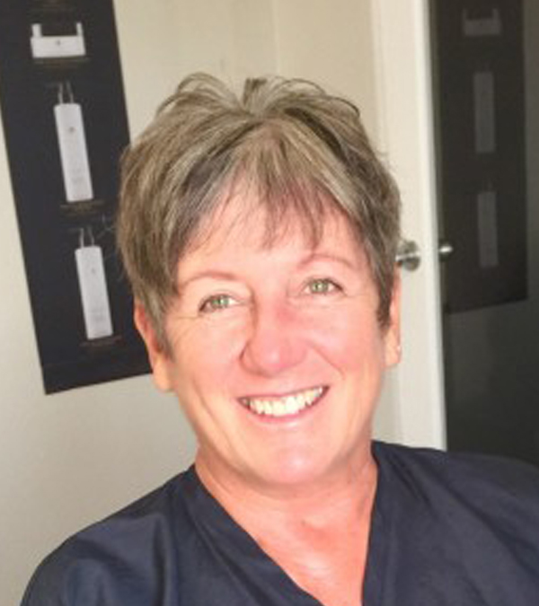 A woman wearing a blue scrub shirt smiles warmly, conveying a sense of professionalism and approachability.