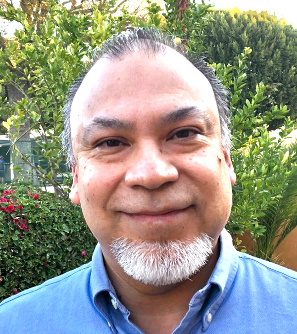 A man with a beard wearing a blue shirt, standing confidently with a neutral expression.